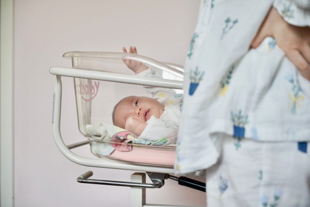 Eine zärtliche Szene: Ein neugeborenes Baby liegt in einem Krankenhausbettchen, und die Mutter steht dicht daneben.