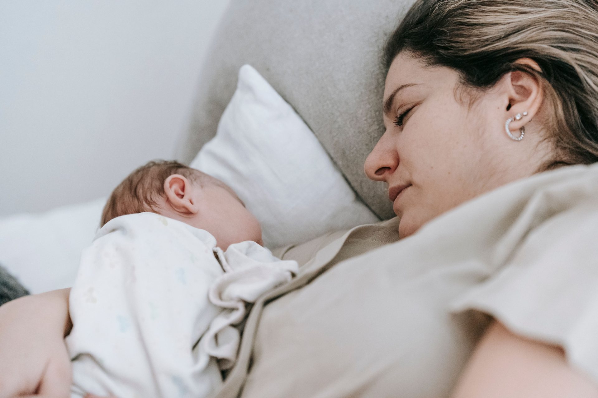 Seitenansicht einer fürsorglichen Mutter mit ihrem süßen, neugeborenen Baby auf dem Arm, das friedlich auf einem bequemen Bett in einem hellen Schlafzimmer zu Hause schläft.