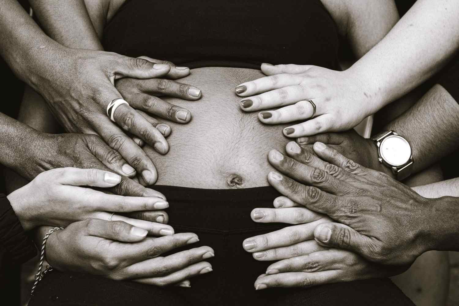 Schwarze und weiße Hände verschiedener ethnischer Zugehörigkeit berühren den nackten Bauch einer gesichtslosen Schwangeren in freudiger Erwartung der Geburt des Babys.
